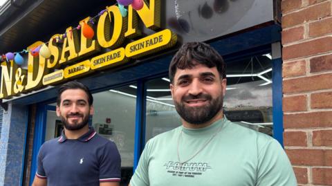 Two men with neat dark beards smile outside a shop front.