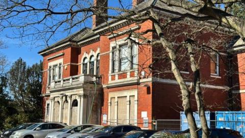 A large, red brick building has its doors and windows boarded up. Cars are parked to the front of the building and a tree with no leaves stands to one side.