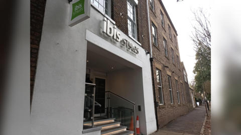 An exterior shot of Ibis Style Hotel in York, and red-brick building with a white plastered facade around the entrance, and the green Ibis style logo sign above. In the entrance, a short flight of stairs can be seen, alongside an accessible lift which has two orange cones placed in front of it. 