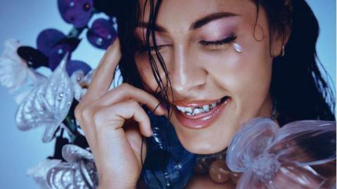 A woman with fake flowers and leaves around her head. She is smiling and has her eyes shut. 