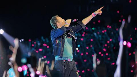A man standing on a stage with a black microphone in his right hand and his left hand – with colourful wristbands on – pointing in the air. He is wearing black cargo-like trousers, a turquoise blue T-shirt and a black jacket with the sleeves folded up to his elbows. He has short curly brown hair. There are pink and purple lights from the crowd blurred in the background.