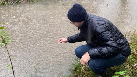 A man bending over a waterway, getting a sample. You can only see the side of his face, and he is wearing a dark coat, dark jeans, blue hat. He is crouched down. 