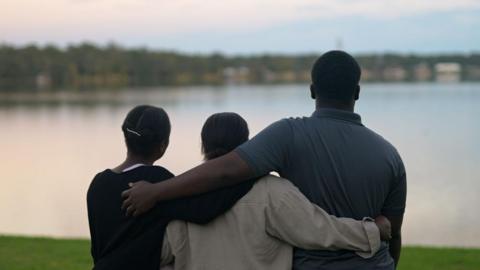Marven, Rochelle and Guerline, pictured from behind, with their arms around each other looking out over a lake. Marwen is wearing a grey, collared T-shirt and is much taller than the two women.