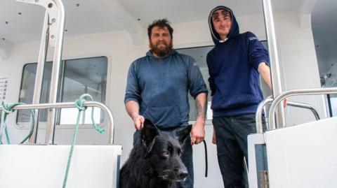 Captain Kimmy Reid with his crew member Aaron, and Bruce the dog. They are standing on a white boat.