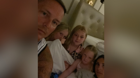 A selfie picture of Kyle and his family. They are sitting on the floor beside a bed.