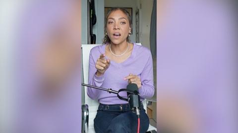 A woman sits in front of a microphone talking to someone across from her. She wears a purple top and dark blue jeans