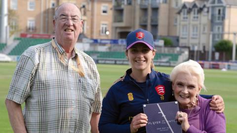 Dave and Pam Letch on the pitch at the County Ground. Dave is standing up while wearing glasses and a chequered shirt. Eva Gray is wearing a navy cricket strip and hat, and has her arm around Pam's shoulders. Pam is wearing a purple outfit and presenting Gray with a player of the match award.