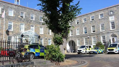 Guernsey Police headquarters. A grey building with three floors and windows on eat floor. There are police vans and cars parked in the car park. There is a tree in the middle of the car park. The sky is blue. 