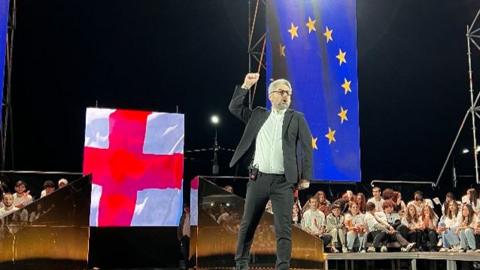 Nika Gvaramia stands on stage pumping his fist in the air, with audience members seated around. The stage is backlit by European Union and Georgian flags