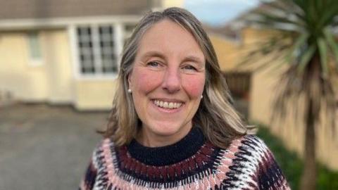 Kerry Sharpe, who has blonde shoulder length hair, and is smiling. There is a house in the background.