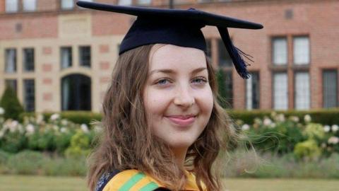 Rachael Moore, who has shoulder length brown hair, smiles at the camera wearing a blue university cap and gown with a yellow and green sash