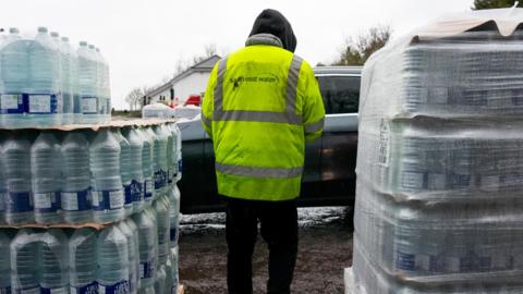 A person in a yellow high-vis South East Water coat. They are standing between two crates of bottled water.