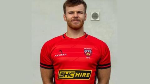 William Bevan. A young man with short hair and a beard wearing a red short-sleeved rugby shirt.