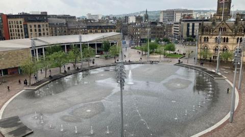 A large public square featuring a circular water feature, with multiple jets spraying water at varying heights. The water gathers to create a mirror effect. Tall light poles surround the fountain, with trees and benches lining the perimeter. People can be seen walking and sitting around the edges of the fountain. In the background, a wider city centre can be seen with a mix of modern and older buildings.