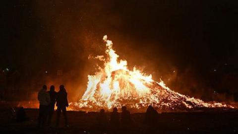A bonfire which is lit on a dark evening with some people standing around it. 