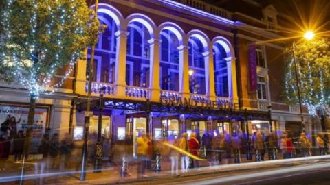 A building with the words Grand Theatre on it is lit up at night with people walking by outside. Two trees with fairy lights on are either side of it, along with a bright street lamp. It is nighttime.
