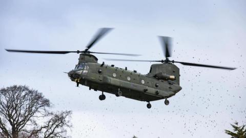 Chinook flying low to the ground just above the tree-line with debris swirling about.