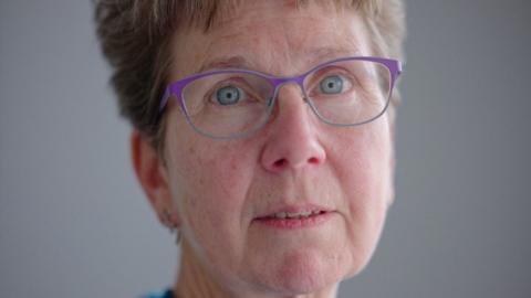 Judith Gibbons has short light brown hair, bright sea-blue eyes and purple rimmed glasses.