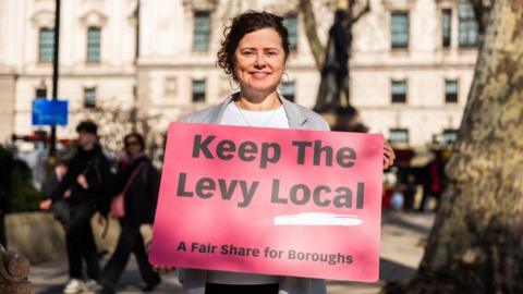 A photo of Claire Holland holding a brightly coloured sign saying 'Keep The Levy Local. A Fair Share for Boroughs'.