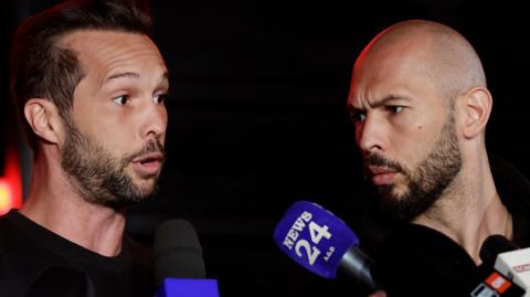 Tristan Tate has a dark beard and dark hair with a receding hairline. He is speaking to the media and two microphones are in the shot. Andrew Tate has a dark beard and a shaved head and is to the right of the image, watching his brother.