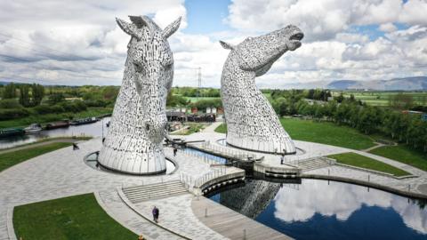 A general view of the Kelpies attraction. The picture is from high in the air. Two horse's heads jut out of the ground. They are silver. There are patches of green grass and water around them.