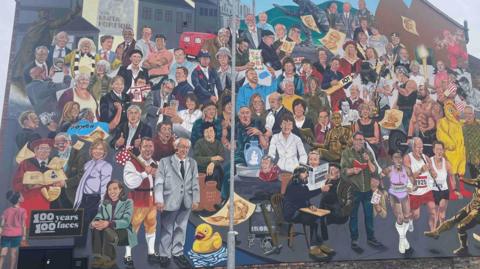 A side of the building completely covered in a painted mural featuring 100 people gathered together with buildings and bottle kilns in the background.
