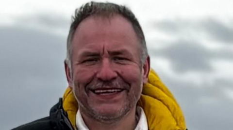 Alan Bolton stands outside where there are grey clouds in the background. Alan has short grey hair and stubble. He is wearing a black and yellow coat. 