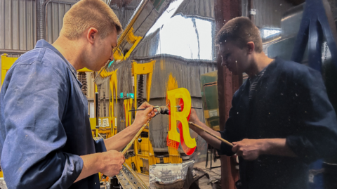 A man painting the letter 'R' onto the side of a locomotive in a works shed