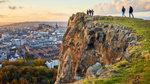 Five people on the edge of a cliff, looking over the centre of Edinburgh. The castle can be seen in the background.