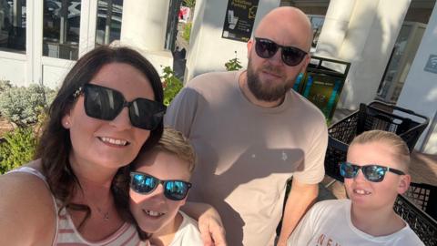 A sunny outdoor selfie shows four people wearing sunglasses gathered close together outside a modern building with white columns and large windows.
Two adults and two children are visible in the foreground, dressed casually in light-coloured summer clothing, with landscaped plants and paved walkways surrounding them.