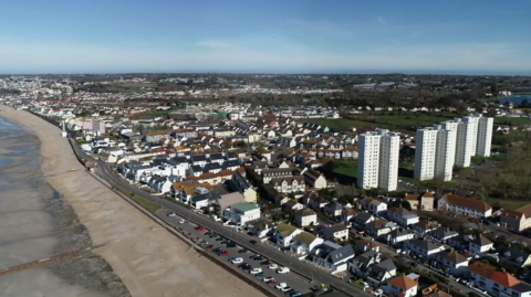An aerial photograph of Jersey's east coast in St Clement. It is a highly residential area, with several homes and four white tower blocks. A two-lane road is along the coast. The beach is on the left of the image, with the tide quite high up the bay.