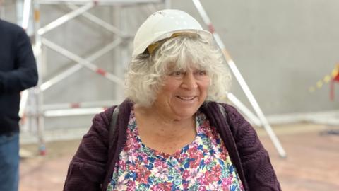 Miriam smiling while being shown around the site. She wears a white hard had perched on top of her grey curly hair, a multi-coloured floral dress and a dark purple textured cardigan. 