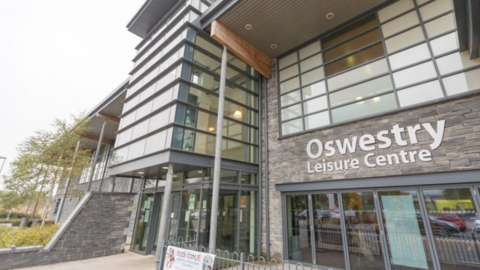 A large building with lots of windows and a stone clad front with the words Oswestry Leisure Centre on the front
