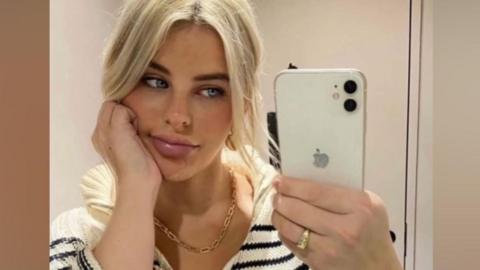 The photo shows Paris Wilson taking a mirror selfie indoors. They are holding a white iPhone with a dual‑camera setup in their right hand, and they are wearing a chunky striped knit top with a zip collar. Their hair appears to be light blonde and tied back loosely. A gold chain necklace is visible around their neck, and they are wearing a gold ring on their right hand. The setting looks like a simple, neutral-toned room, possibly a bedroom or changing area.