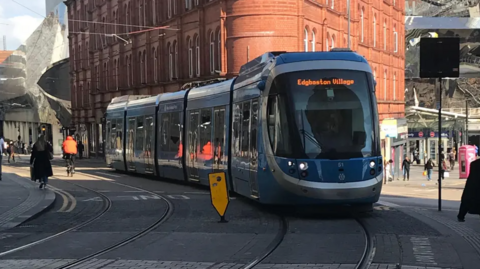 A tram in Birmingham city centre heading to Edgbaston. It is on track near New Street with pedestrians walking nearby.