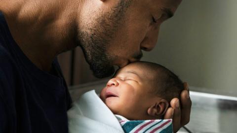 A father kisses a new born baby on the head. The baby is wrapped in a striped blanket.