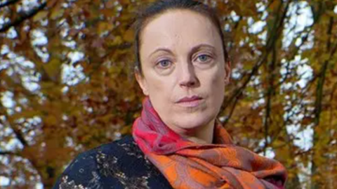 A woman with tied-back brown hair, an orange and pink scarf and a dark blue top in front of trees