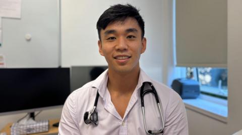 Headshot of Dr Brian Wang in a clinical office. He is wearing a purple shirt with a stethoscope around his shoulders