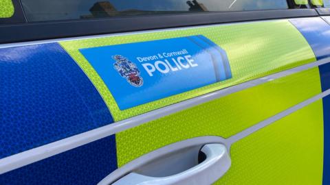 The side of a blue and yellow police vehicle. 'Devon and Cornwall Police' is written in a blue box on one of the doors of the car.