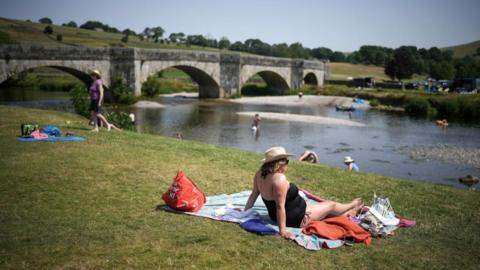 A woman in a black bathing suit sits on a towel on a patch of grass with a stone bridge and water in the distance.