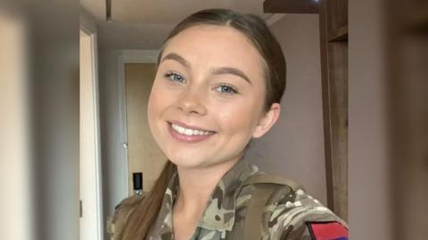 A selfie of Jaysley Beck wearing her military uniform, with her long brown hair slicked back into a low ponytail. She has blue eyes and is smiling at the camera.