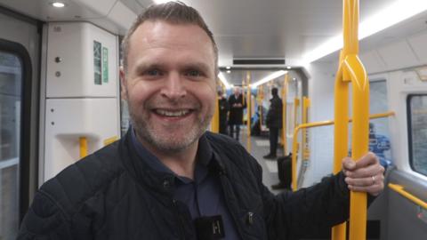 Mark Bryant, 46, stood smiling at the camera on board a new tyne and wear metro train. He is wearing a blue shirt and black jacket, and is holding onto a yellow railing.