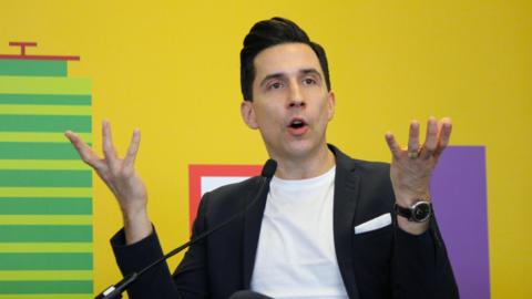 Russell Kane, a man wearing a white t-shirt and a black jacket is gesticulating with his hands as he talks into a microphone. He is pictured against a brightly coloured backdrop which is yellow and has additional block colour elements.