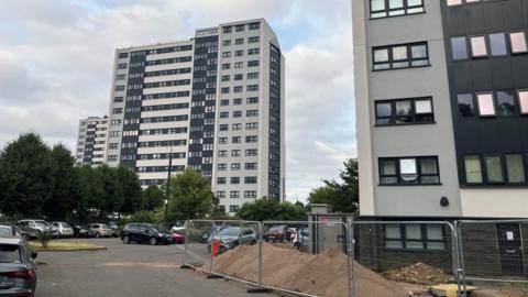 A row of three 15-storey tower blocks, with one fully visible in the middle of the shot, the side of one block in the foreground, and the third visible in the distance behind the middle one. All three are rendered in light grey with black panels and windows.   
