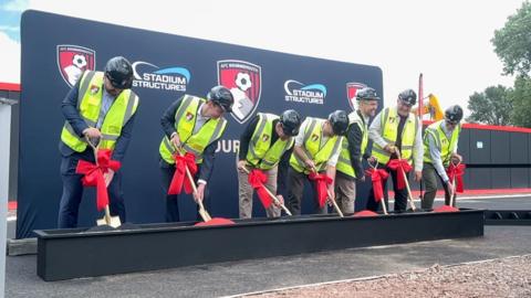 A group of seven men are all lined up wearing suits and yellow high vis vests. They are each holding a golden shovel with a red bow tied around the stems as they dig 'red' soil from a planter.