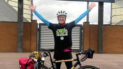 Kate Strong standing outside the football stadium alongside her bike with her arms in the air
