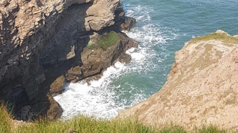 A grassy cliff drops steeply to deep blue water where waves crash against jagged rocks.