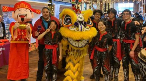 A bright yellow Chinese lion made of fury material surrounded by five dancers wearing black and red outfits. Also a bright red God Of Fortune mascot at the side holding a golden lamp.