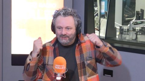 Michael Sheen who has grey hair and a grey beard, sat in a radio studio behind an orange microphone with black headphones on his head. He is wearing an orange and black chequered jacket
