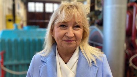 A woman with blonde shoulder length hair wearing a light blue jacket and a white scarf.